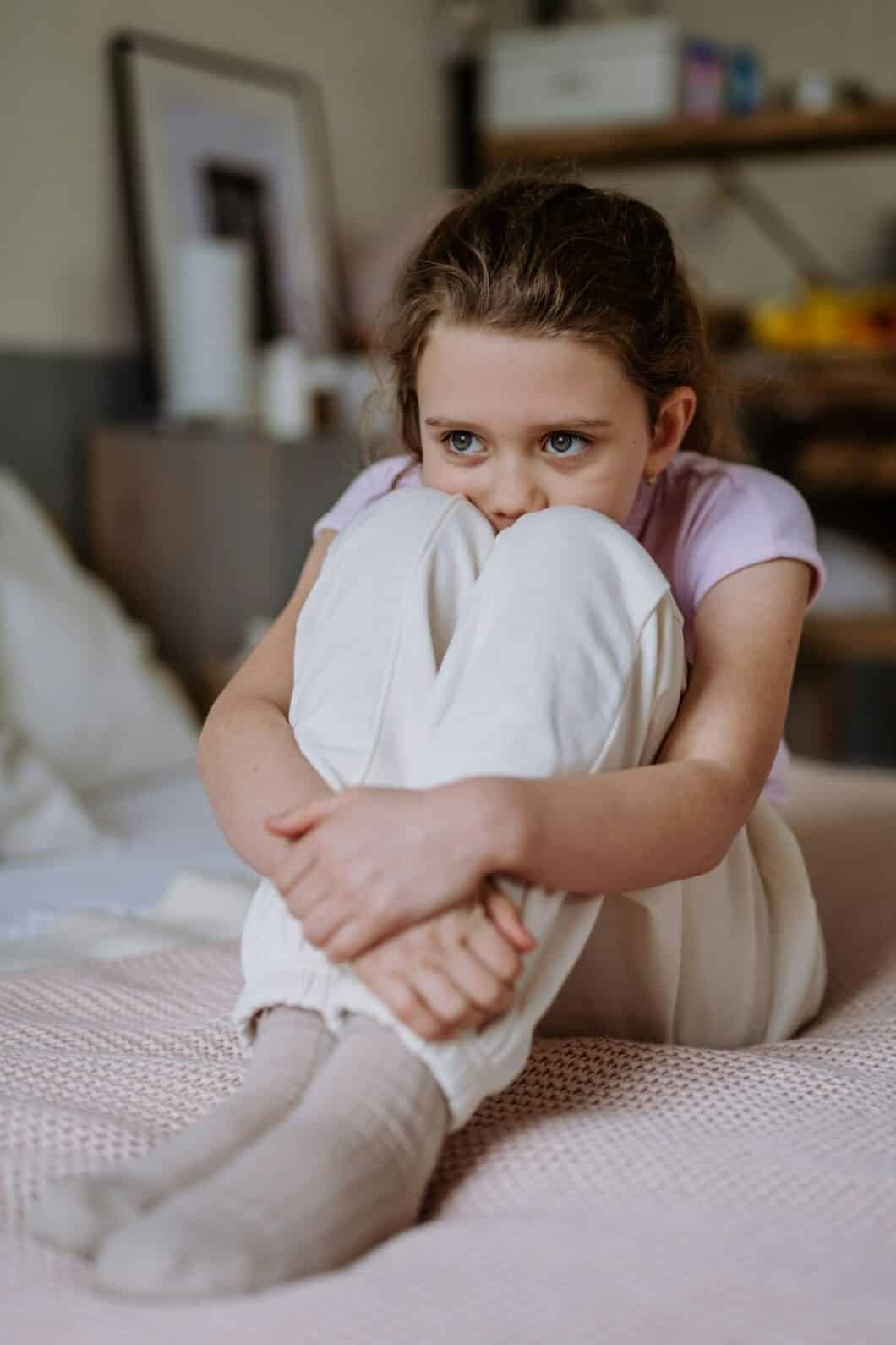 Little sad girl sitting alone on a bed.